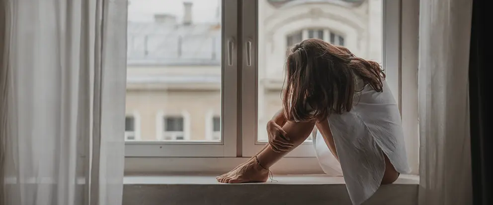 Mujer mirando por la ventana, manifestando estrés debido al atraso menstrual. Solución inmediata.