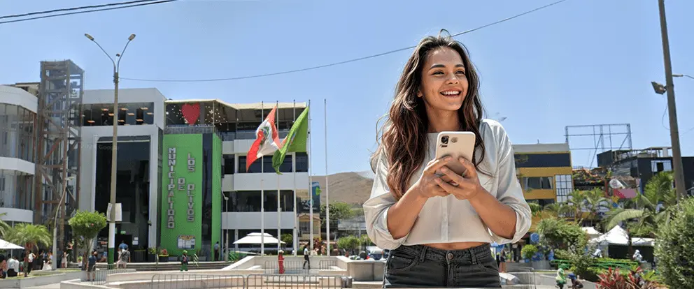 Mujer joven utilizando su teléfono móvil frente a la municipalidad de Los Olivos, buscando información sobre tratamientos para el atraso menstrual.